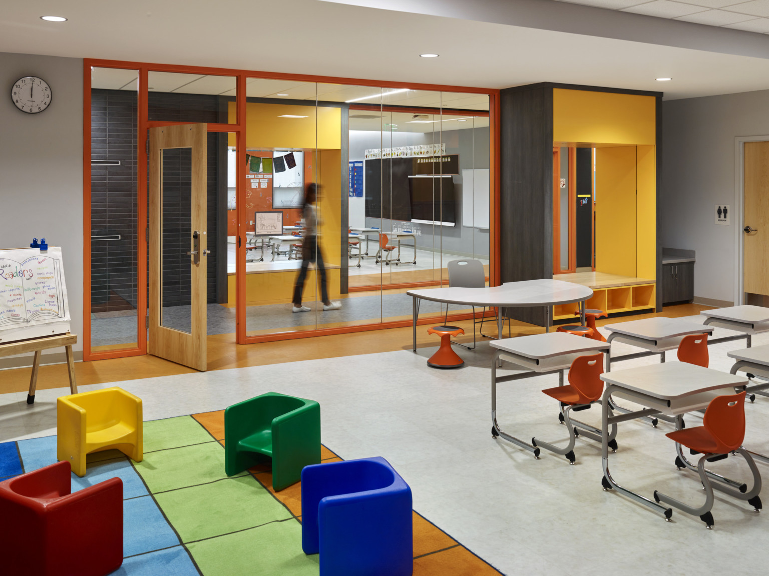Classroom with glass wall to hallway. Flexible desks face table at front. Stools on padded carpet. Yellow booth with cubbies