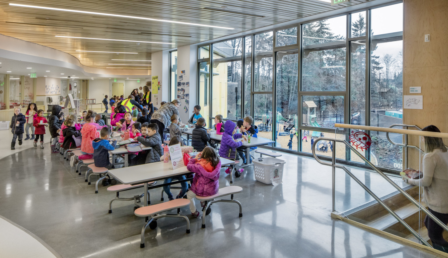 A lunch room with floor to ceiling windows with views of the playground. Above, a curved wood ceiling detail A lunch room with floor to ceiling windows with views of the playground. Above, a curved wood ceiling detail