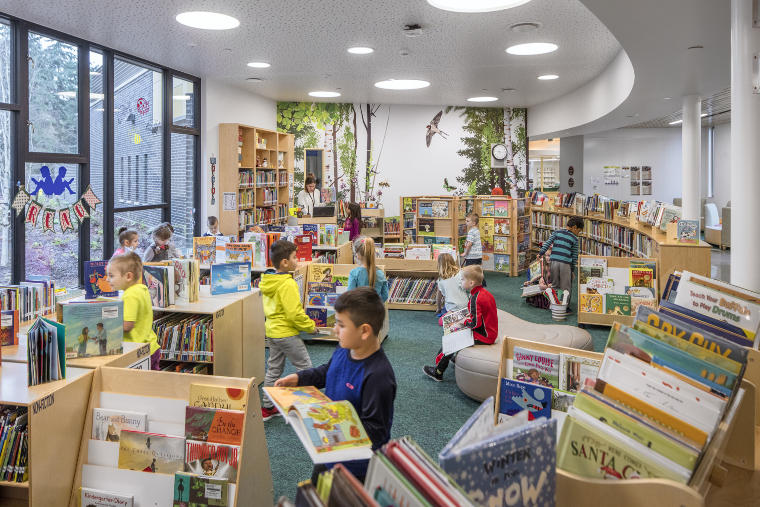 A library with children's bookshelves and seating. Floor to ceiling windows, left. A mural on far wall of trees and birds