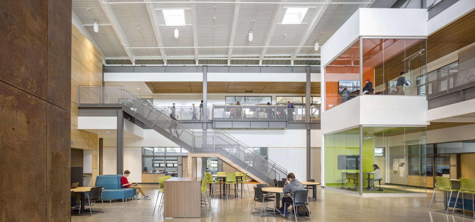 Atrium in Discovery High School with double height ceiling and 2 levels of colorful glass learning pods. Grey stairs, left