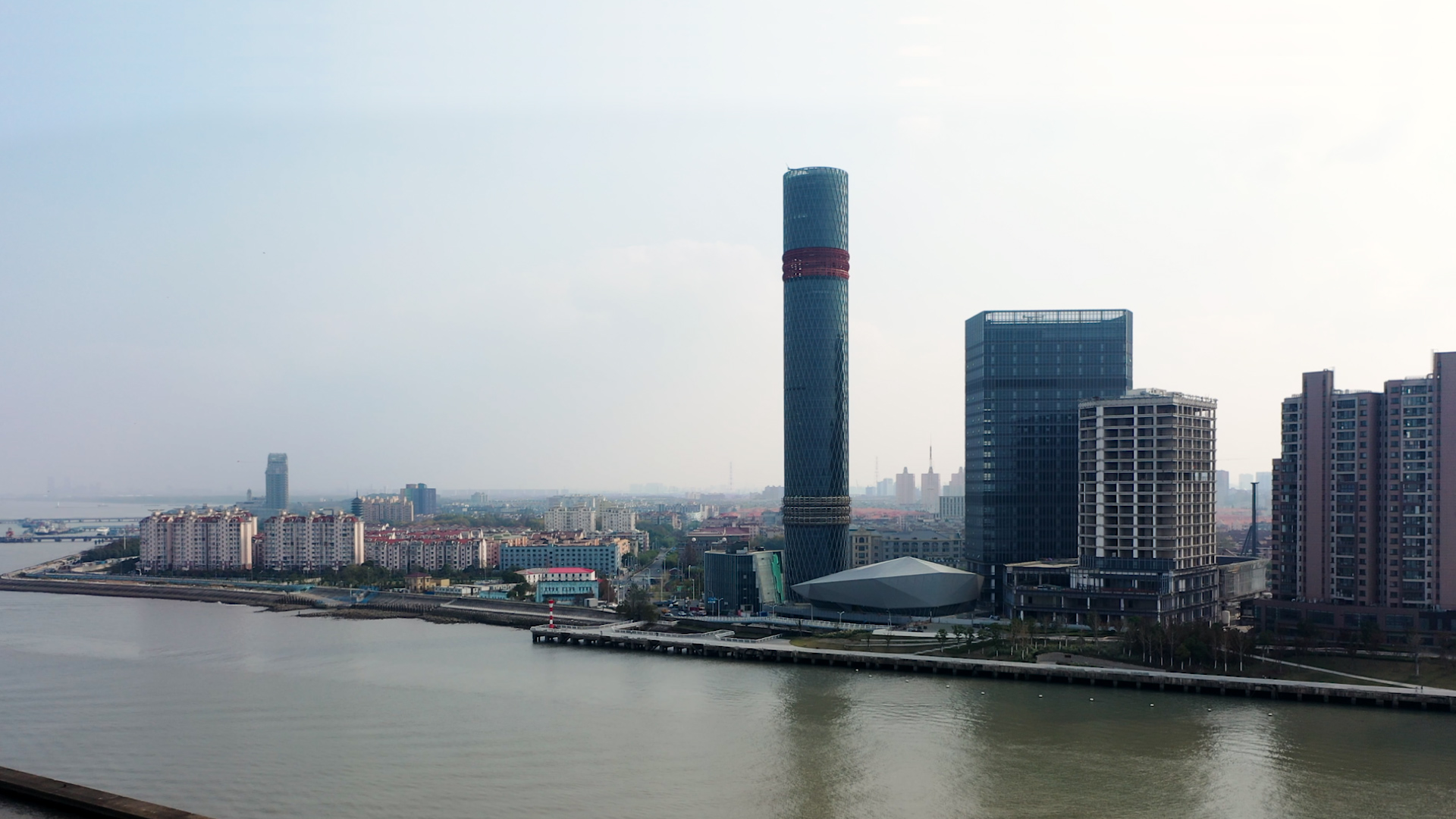 Mixed use campus seen from the water. Tall highrise tower in the middle of the frame with blue glass facade and red design band in Baoshan China