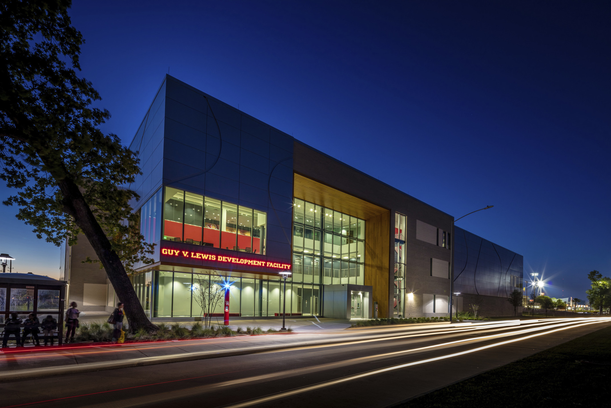 Guy V. Lewis Development Facility illuminated at night with red sign left of triple height glass entrance with wood accent Guy V. Lewis Development Facility illuminated at night with red sign left of triple height glass entrance with wood accent