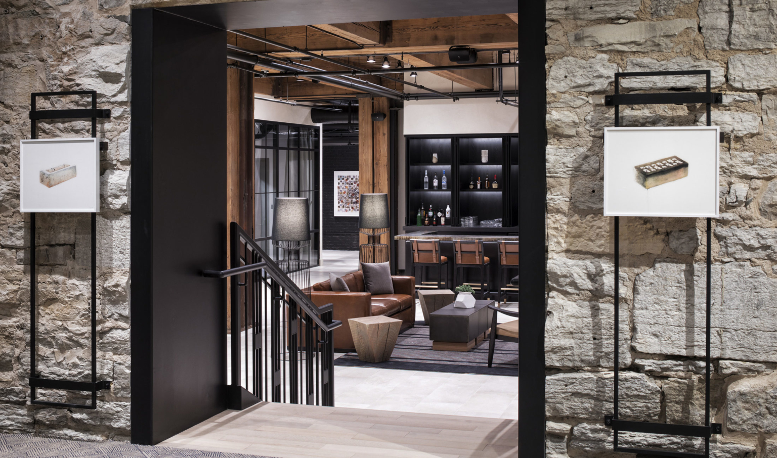 Stone wall with black doorframe leading to short stairwell. Below, seating area with exposed wood beams and columns