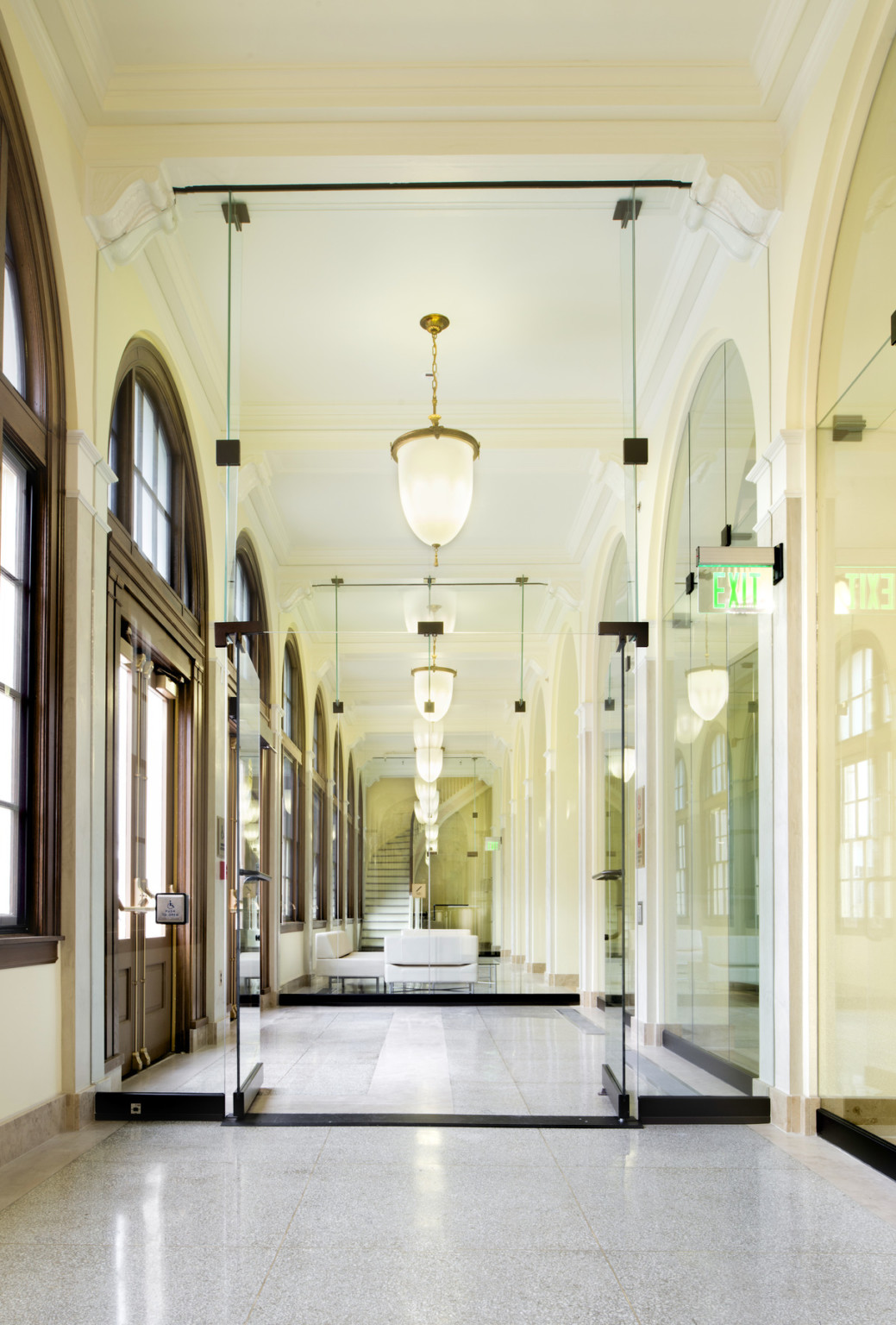 A hallway lined with arched windows and mirrors and central pendant lights leading to a curved stairwell