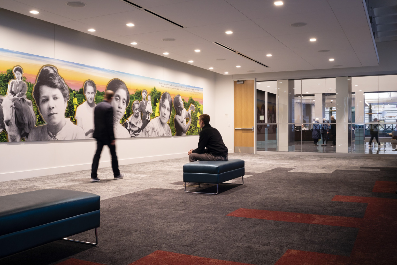First floor event space with geometric carpet, a man sits looking at a large colorful mural