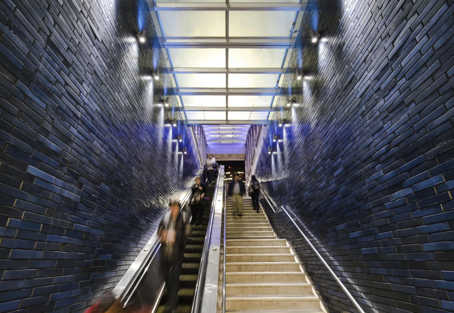 An illuminated glass panel canopy ceiling over subterranean escalator (left) and stairs (right) with tiled walls