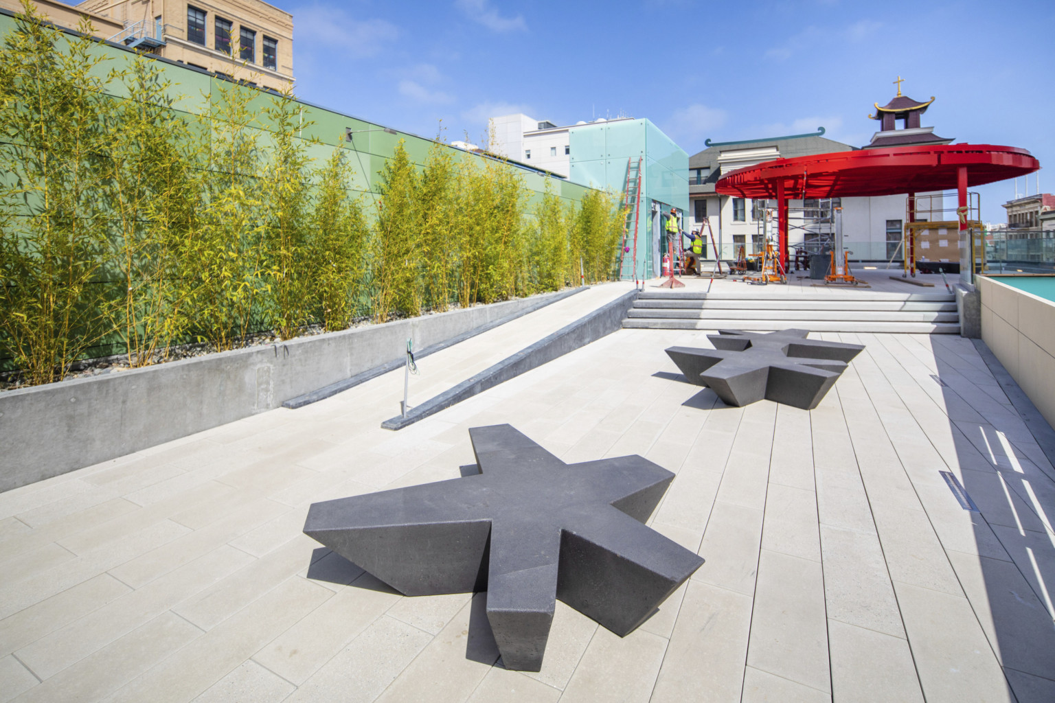 Organic shaped benches in walkway with ramp and steps to rooftop plaza with red canopy. Trees grow along green tile left wall