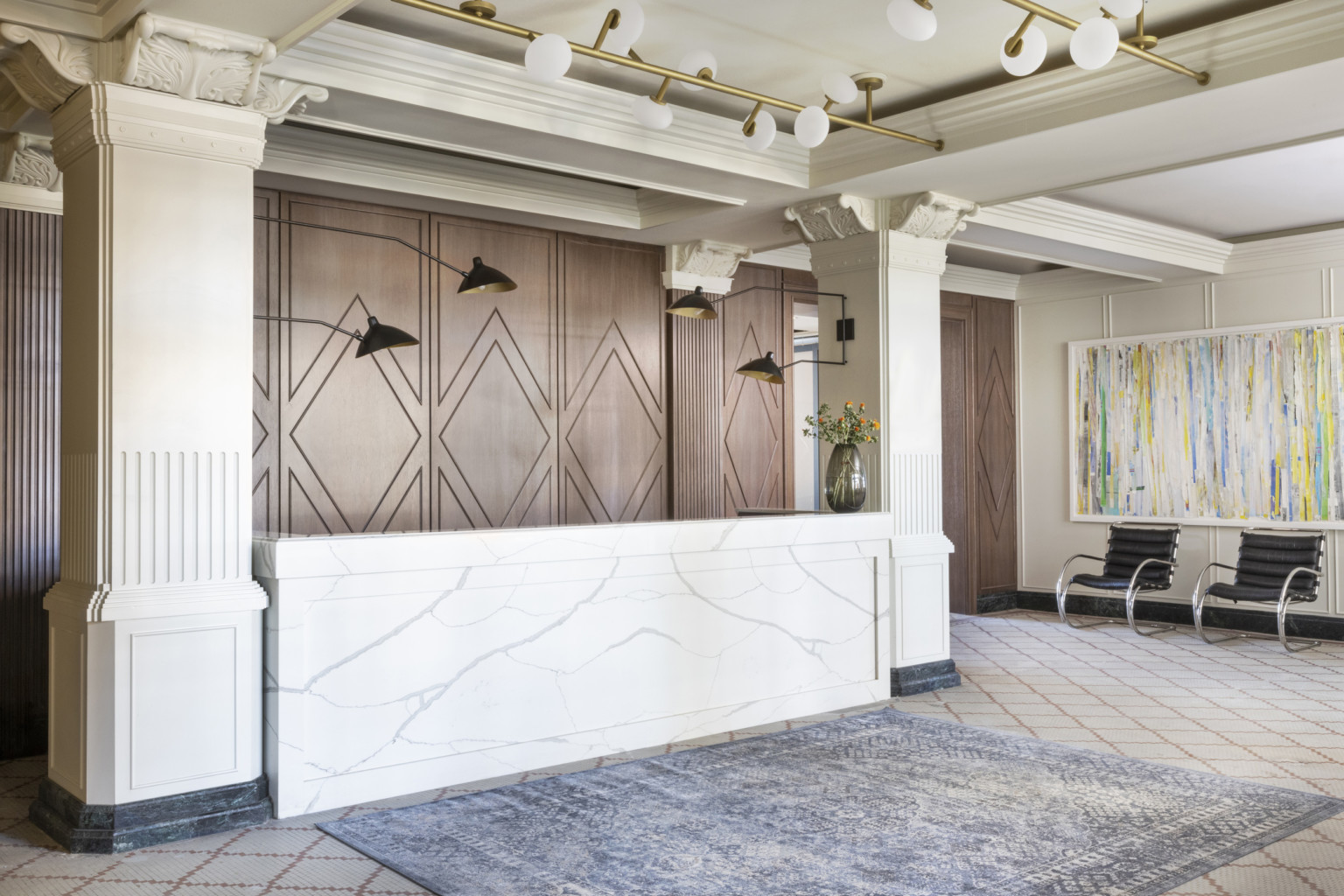 Lobby with white marble reception desk between Corinthian columns in front of geometric wood wall. Blue rug in front of desk