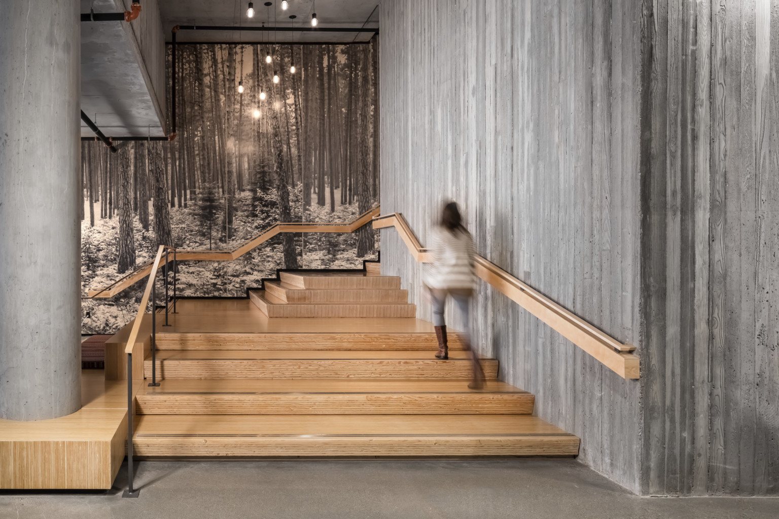 woman walks up wooden stairs surrounded by raw concrete walls. Black and white mural, front of a forest. Angled railing woman walks up wooden stairs surrounded by raw concrete walls. Black and white mural, front of a forest. Angled railing