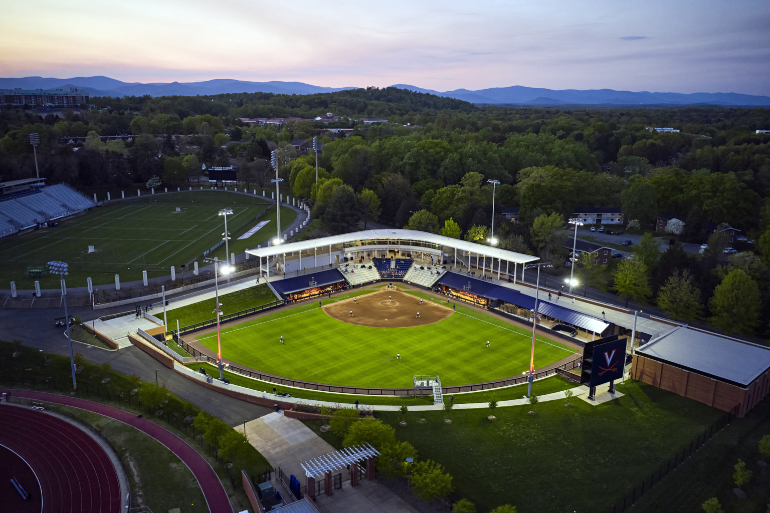 Aerial view of softball stadium at dusk lit up but lights and surrounded by lush trees