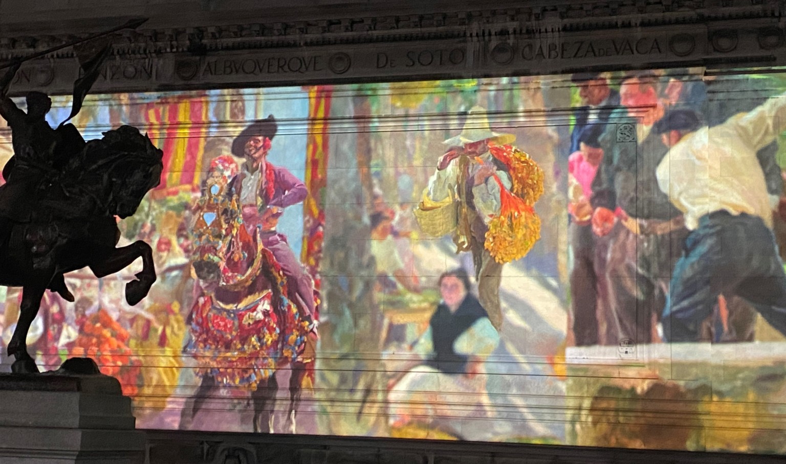 Colorful paintings projected on the exterior wall of Beaux-Arts style building at night. Sculpture of knight and horse, left