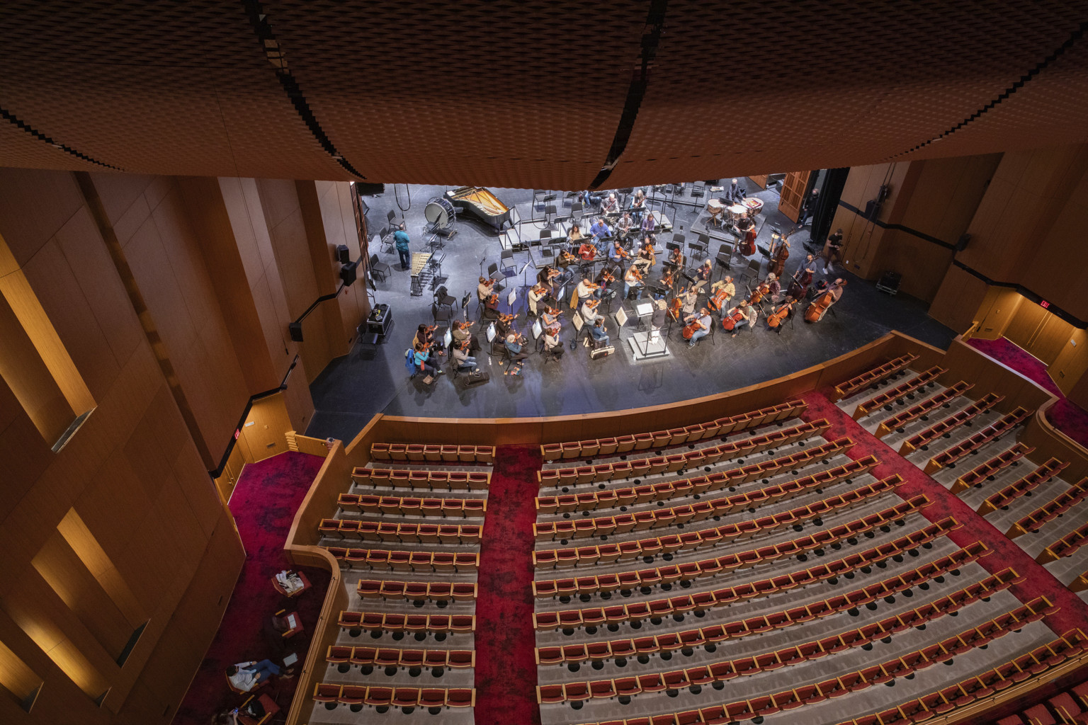 Looking down from a theater balcony showing a wood panel theater, red carpeting, and up lighting while a symphony plays on a large black stage