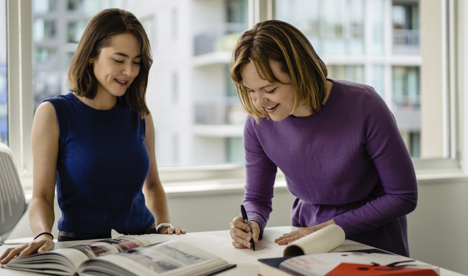 2 women stand at table with books opened. Woman, in purple shirt to right, writes on a pad with a marker 2 women stand at table with books opened. Woman, in purple shirt to right, writes on a pad with a marker