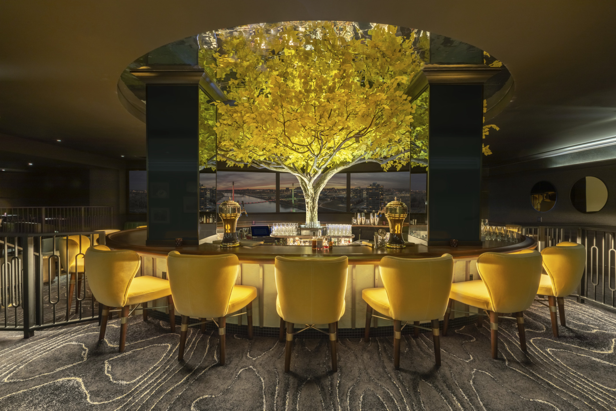 an illuminated yellow tree in the midst of in the round high top bar with yellow seats. Tree rises above round ceiling cutout