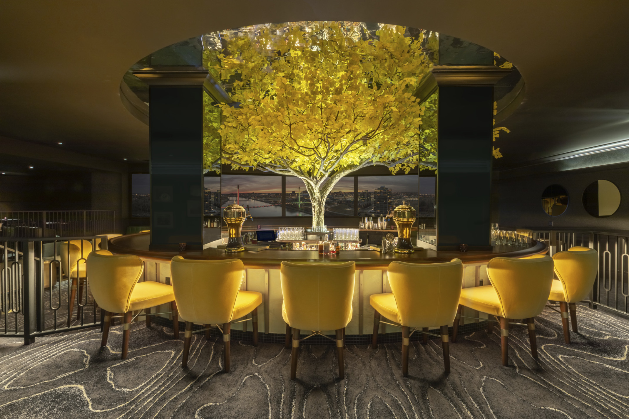 an illuminated yellow tree in the midst of in the round high top bar with yellow seats. Tree rises above round ceiling cutout an illuminated yellow tree in the midst of in the round high top bar with yellow seats. Tree rises above round ceiling cutout
