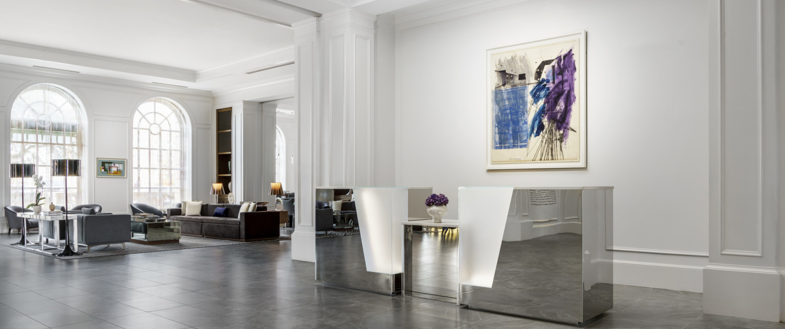 Metallic reception desk with illuminated detail in white hotel lobby with large arch window, and seating area left