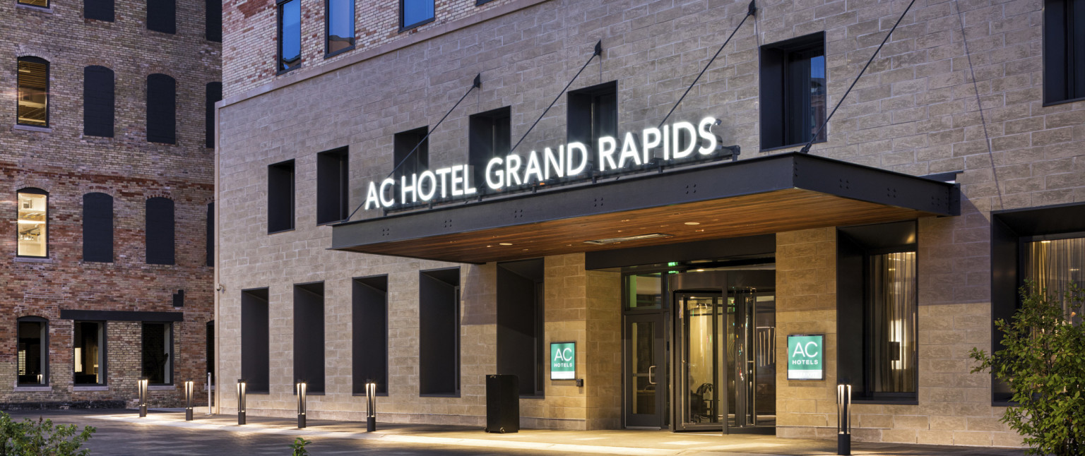 Exterior entry to the AC Hotel Grand Rapids with illuminated sign on awning. Stone lower levels beneath brick upper stories