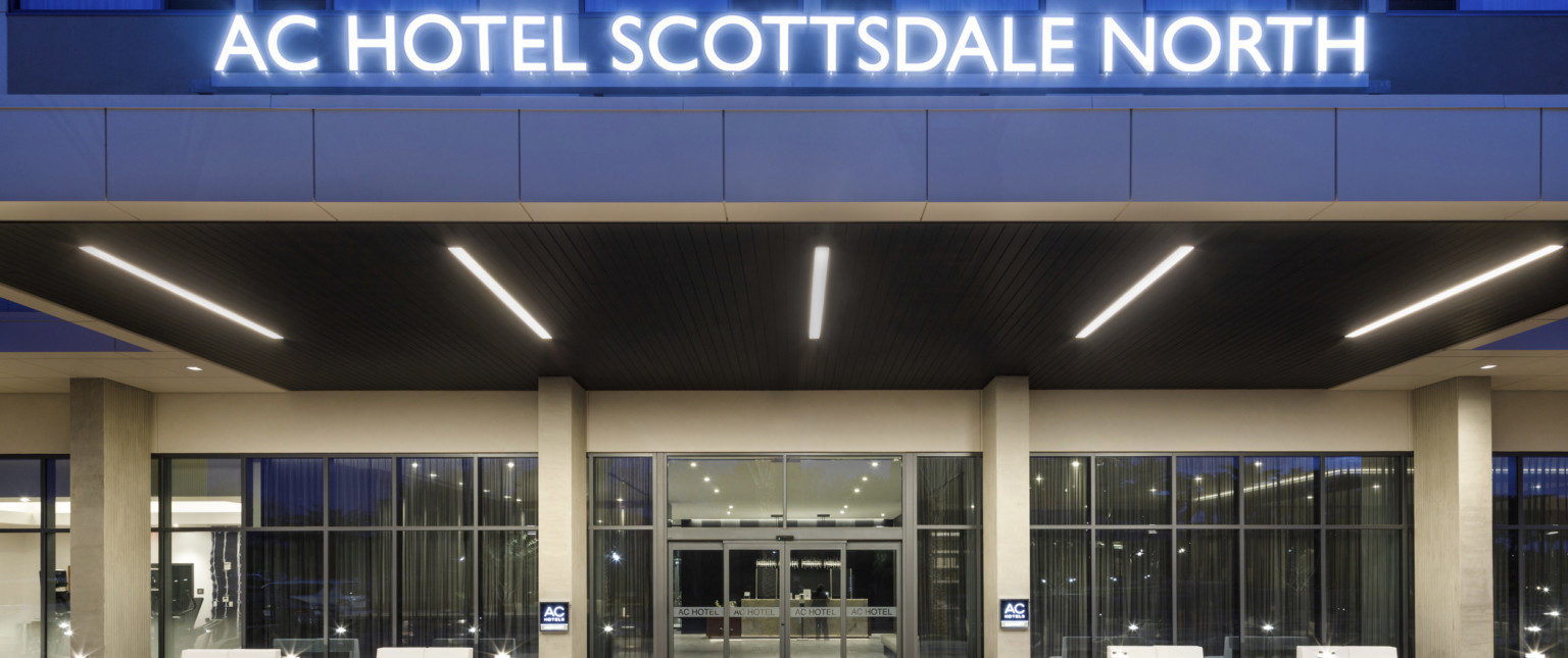 Close up of hotel entry with signage illuminated in blue above. Stone overhang with wood panels under and recessed rod lights