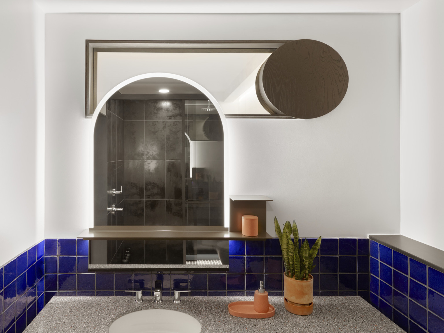White bathroom with blue backsplash around grey counter with sink. Arching mirror centered over sink below geometric shelf