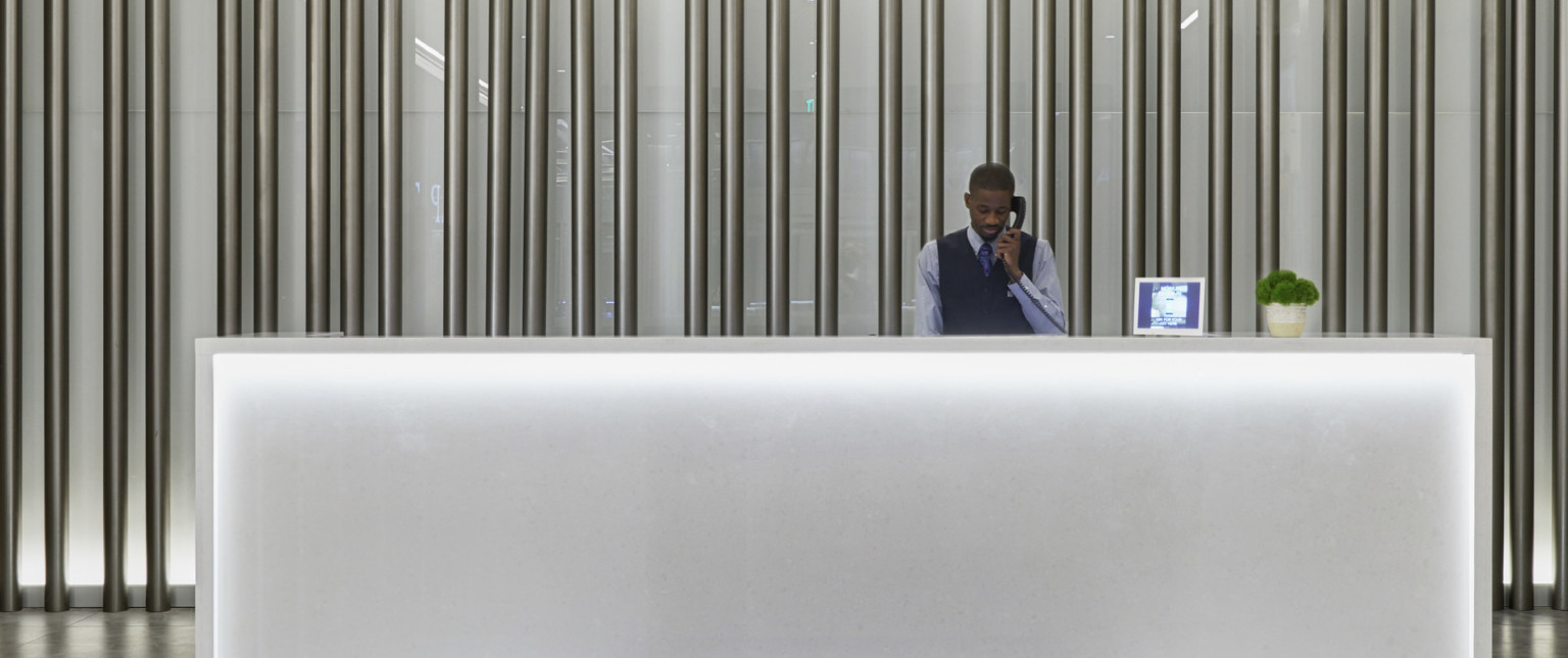White illuminated reception desk with man answering phone. White back wall with silver tubes running parallel