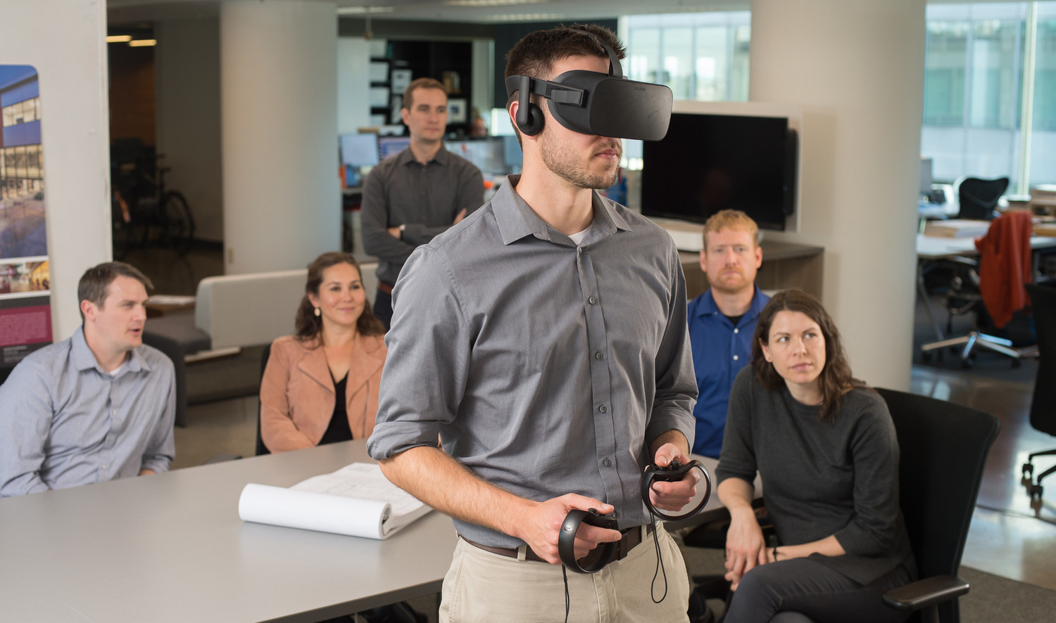 Man uses VR headset and controls in front of table with 5 people in white office, desks back right. Pad of paper on table