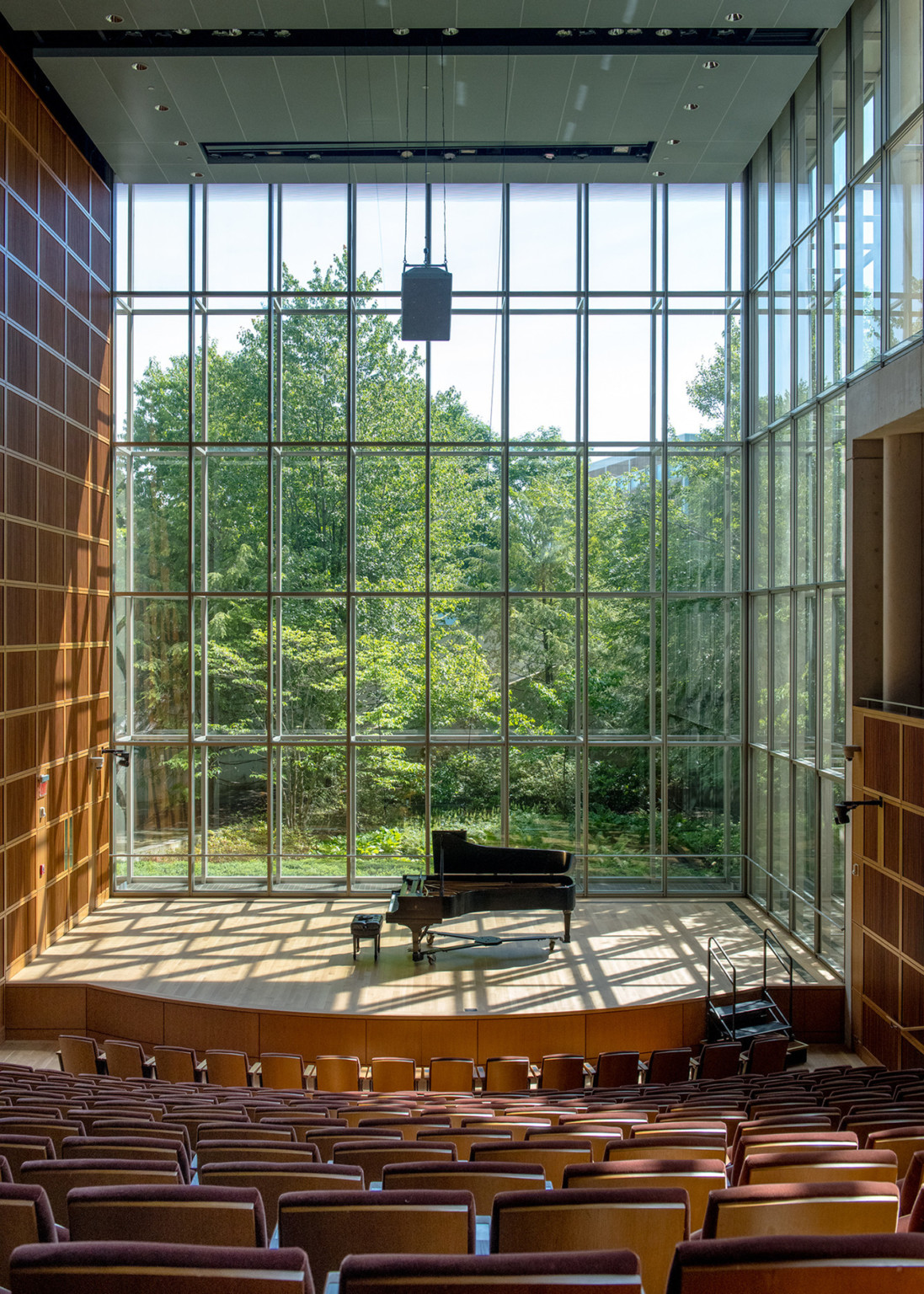 a piano on a recital stage against a floor to ceiling wall of gridded windows flanked by wooden paneled acoustic walls