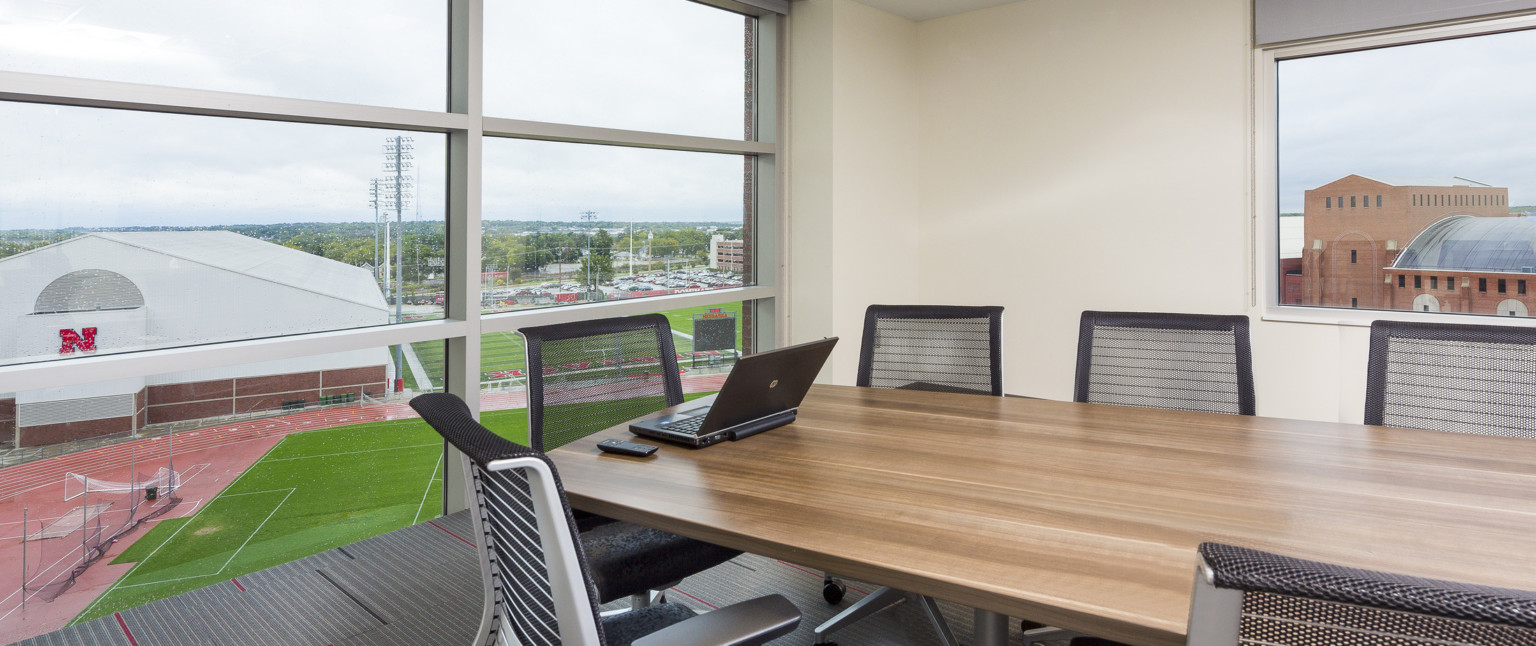 Corner conference room with wood table and black chairs. Floor to ceiling windows with white seams, left, view fields