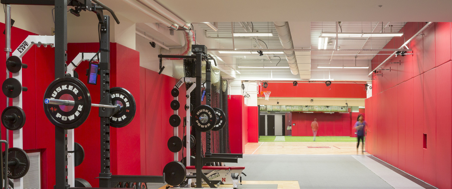 Exercise equipment in white room with red wall padding and exposed ductwork. Beyond, sections of hard wood and turf floor