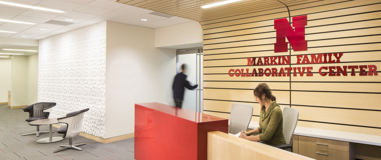 Wood reception desk with red partition in front of wood panel wrap accent wall with Markin Family Collaborative Center sign