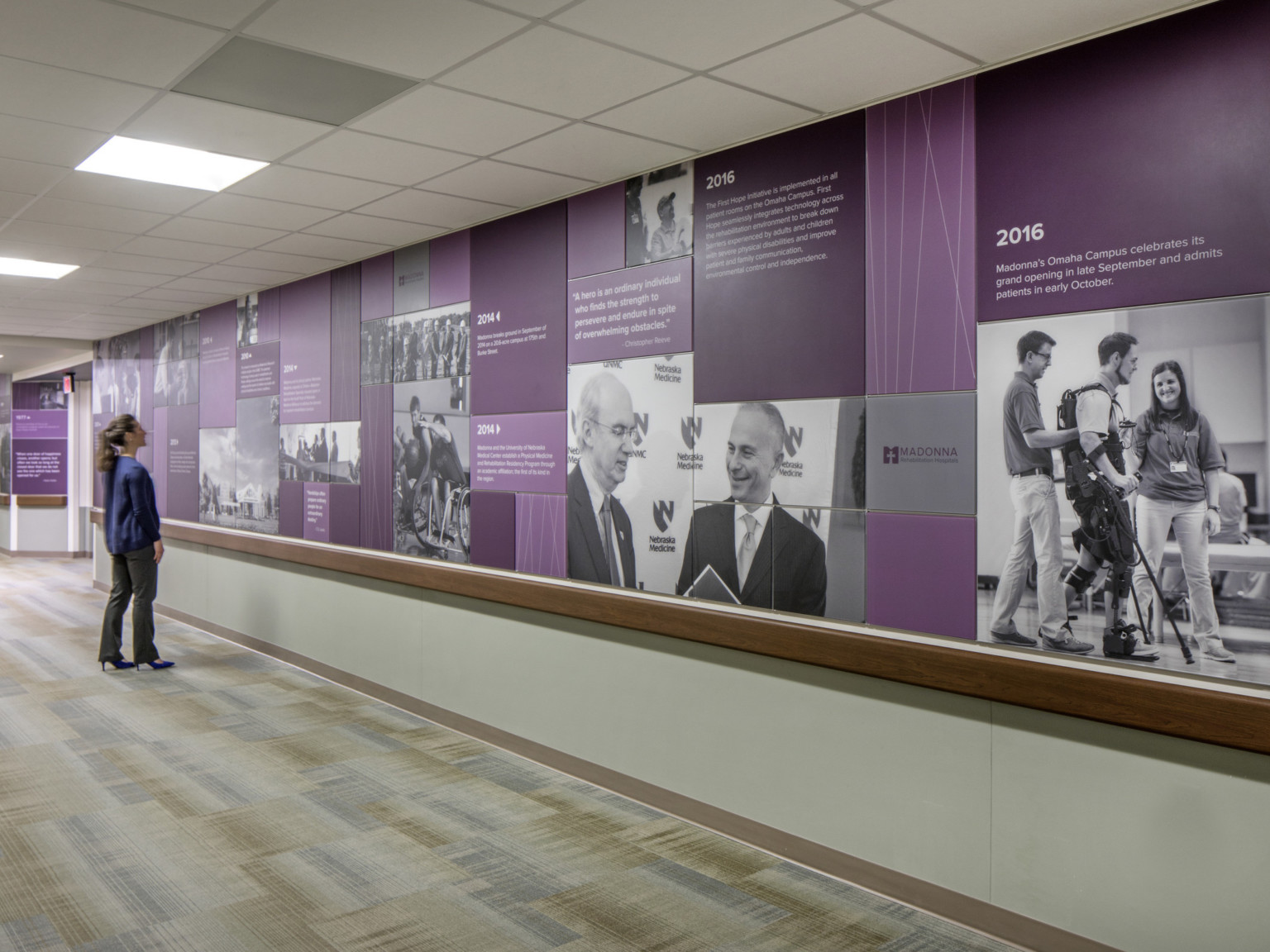 Carpeted hallway with mural on top half on wall. Mural shows timeline on purple panels among black and white photos