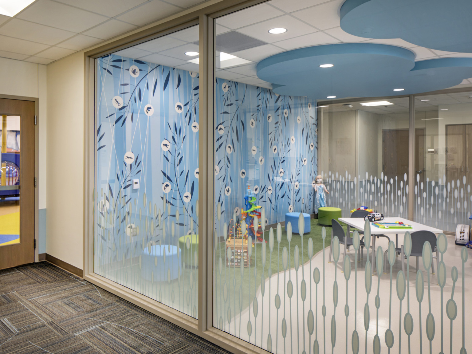Hall looks into glass walled playroom blue abstract floral mural on back wall, and blue cloud shaped drop ceiling detail