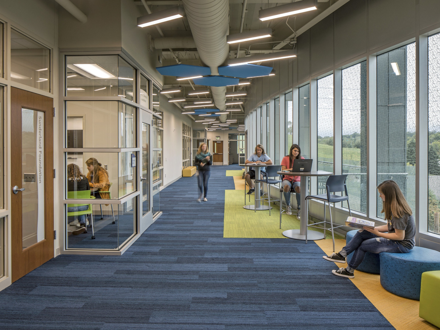 Hallway with blue carpet and blue pentagons hanging overhead. Large windows on the right and glass classrooms to the left