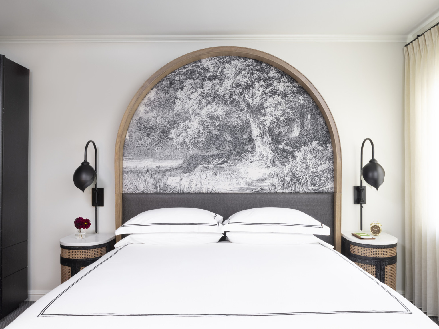 Bed with white comforter framed by wooden arch over bed frame with black and white tree mural. Bedside tables at each side