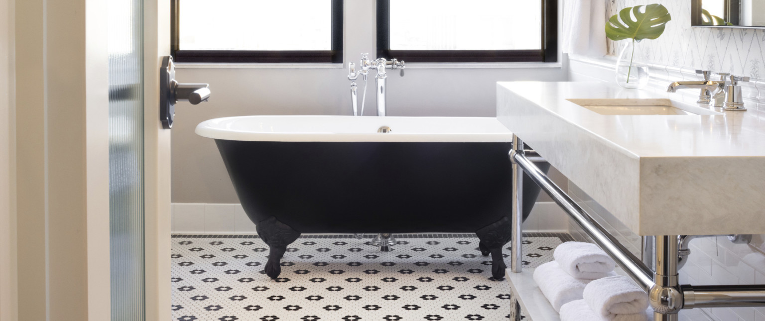 Black clawfoot bath tub with white interior and silver faucet at center in white room with black and white tile, marble sink