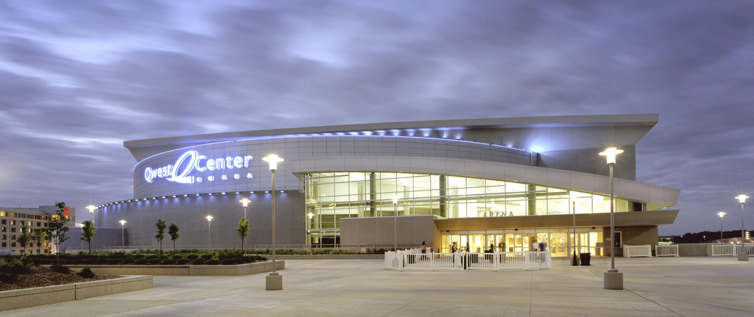 Arena entry illuminated at night with lit Qwest Center sign to left. Planters with trees and shrubs in front plaza