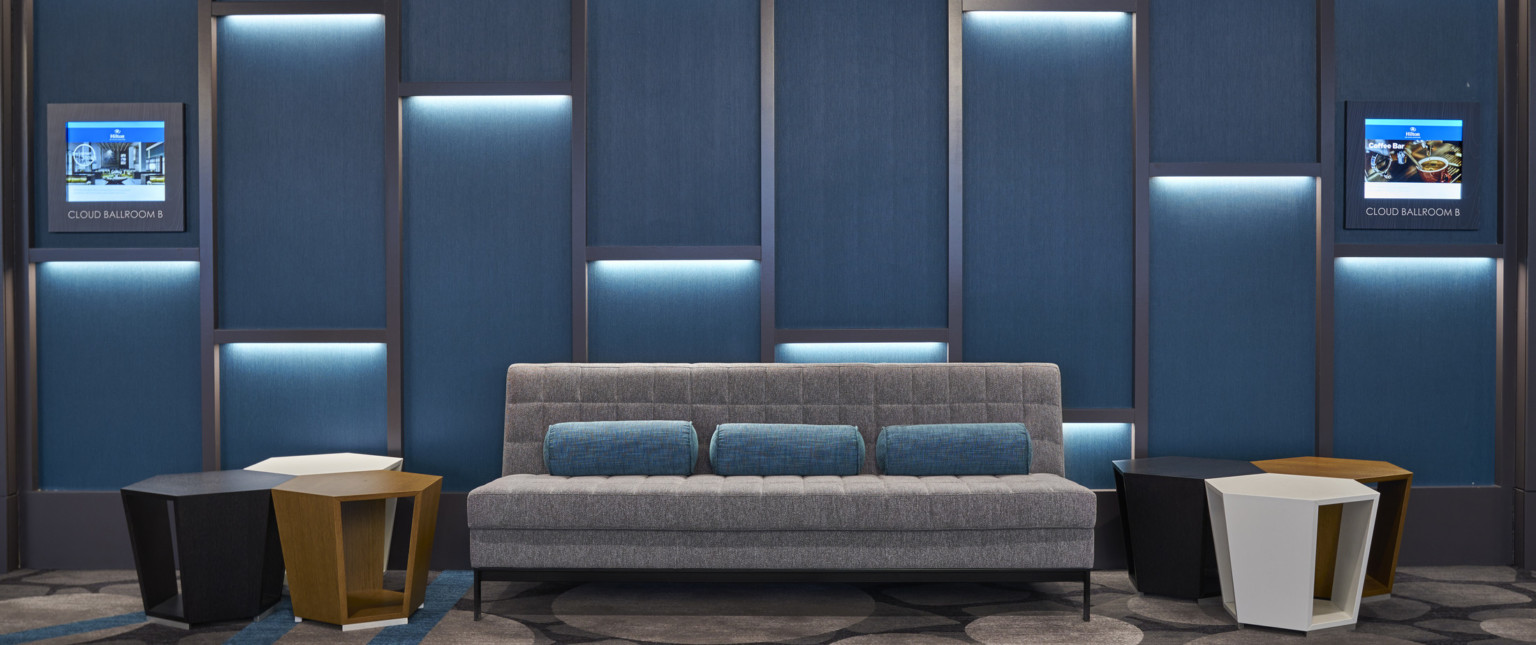 Grey couch with geometric side tables against blue wall panels with silver repeating angled texture with recessed lighting
