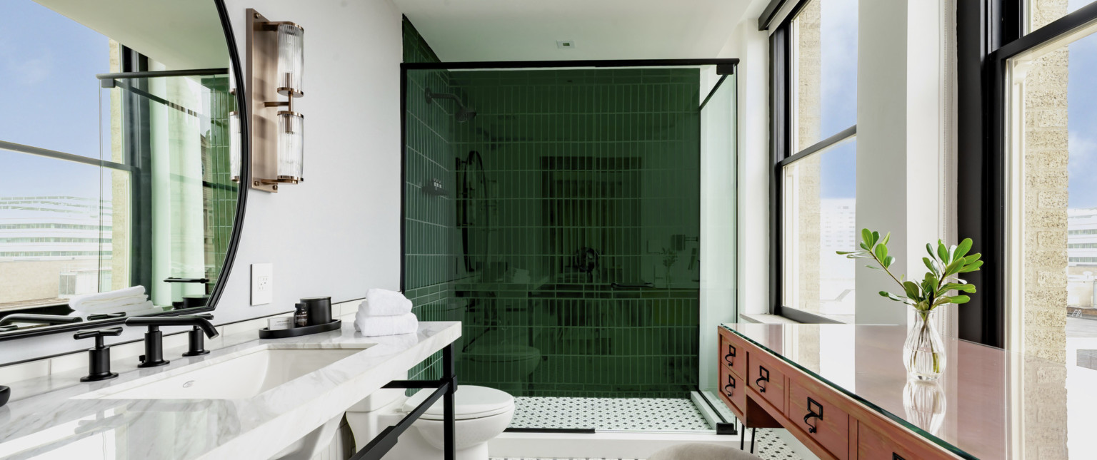 Bathroom with green tiled shower, toilet, and sink left. Right are 2 large windows in front of table and stool