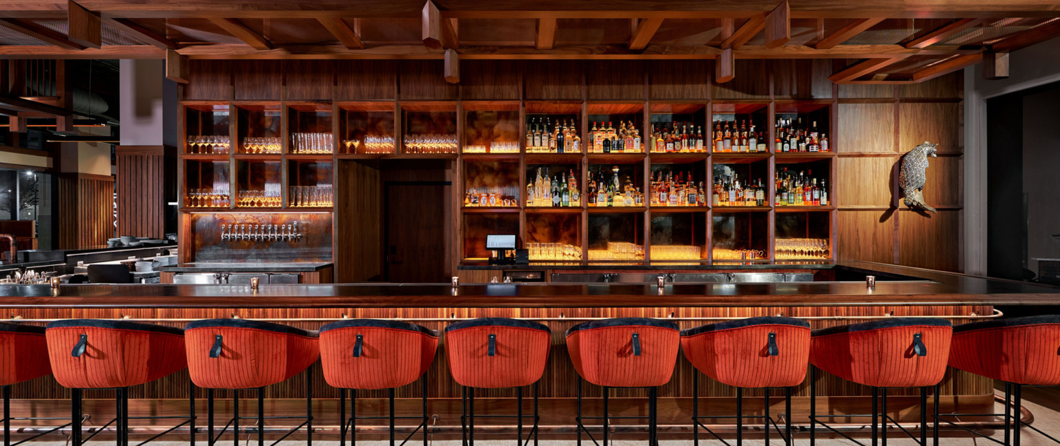 Large wood bar with built in shelves along the back. Wood overhang over back of bar, and stools with red and black upholstery