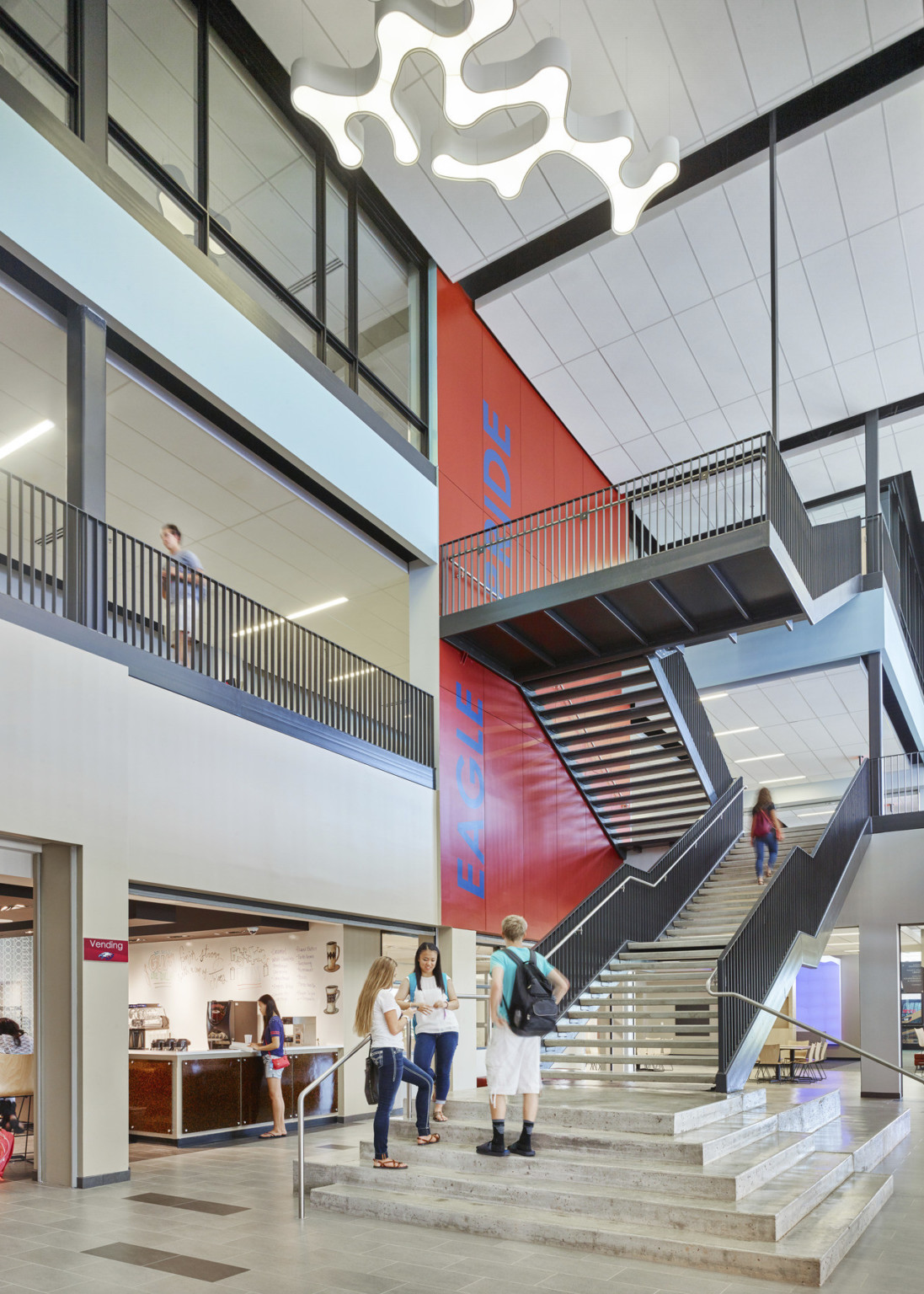 Wood floating stairs with black railing in white 3 story atrium. Orange accent mural to left of stairs reads Eagle Pride