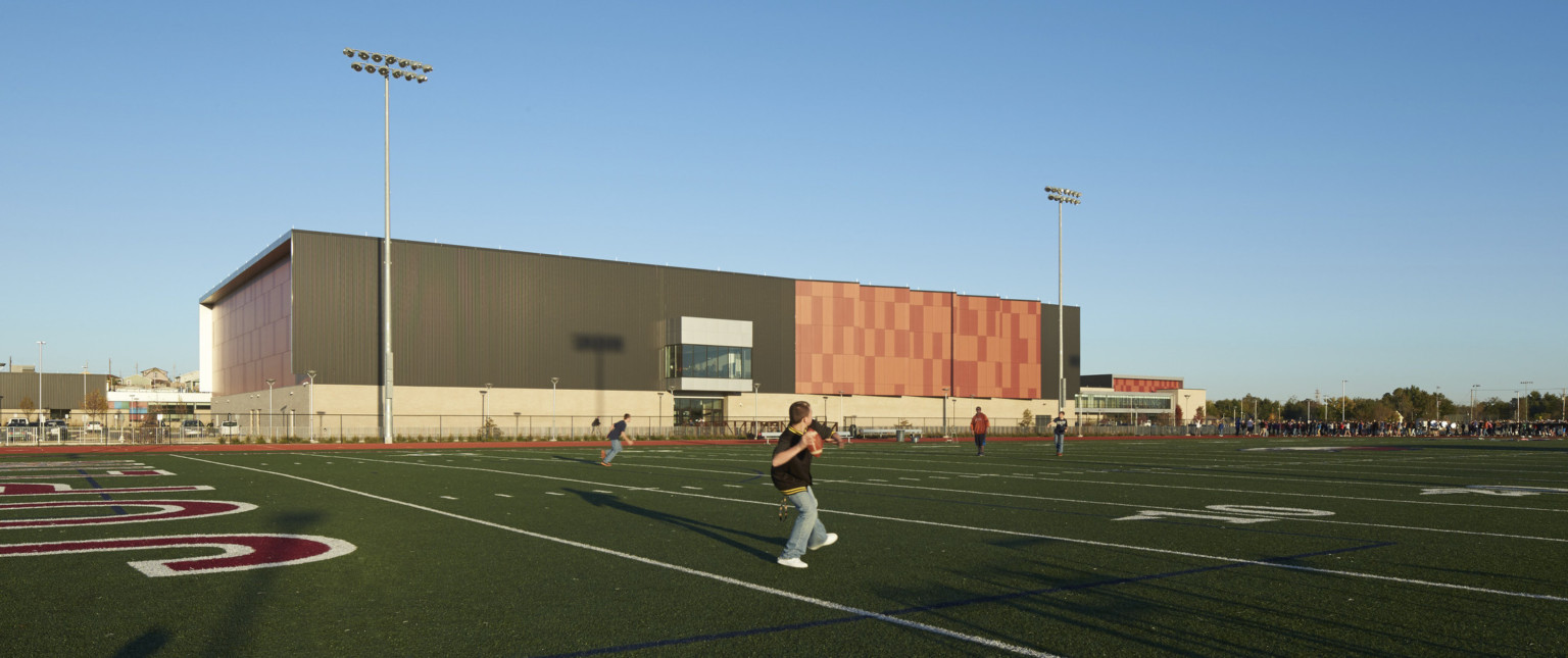 Football field with stadium lights to back of orange panel building with black wrapped facade and orange serrated accents