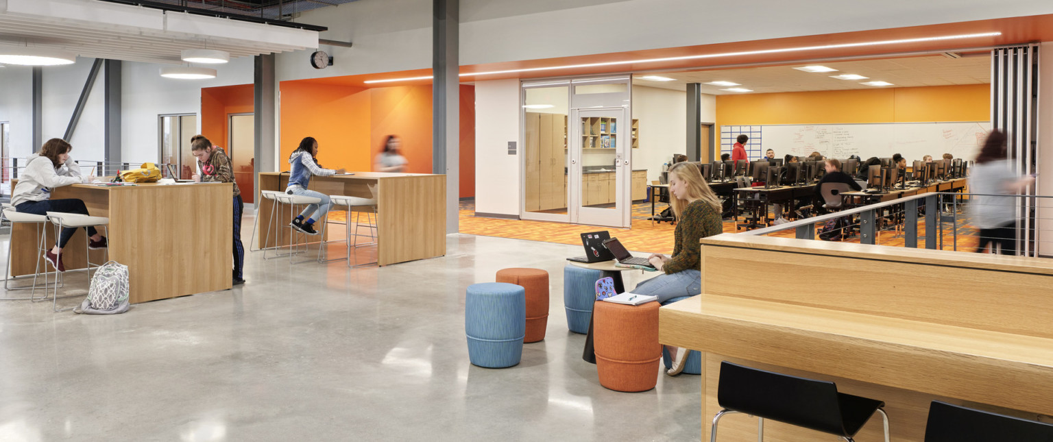 Flexible seating area in hallway with orange accent walls to right. Computer lab with opened flexible wall to hallway
