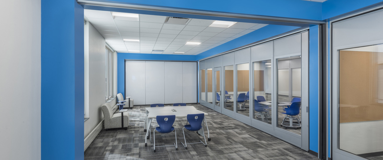 Large whiteboard in blue room with mixed seating. Silver and glass flexible wall, right, with flexible desks in next room