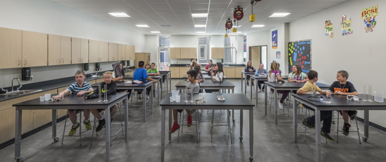 White science lab with light wood cabinets, black counters and rows of lab tables. Extension cords above, windows at back