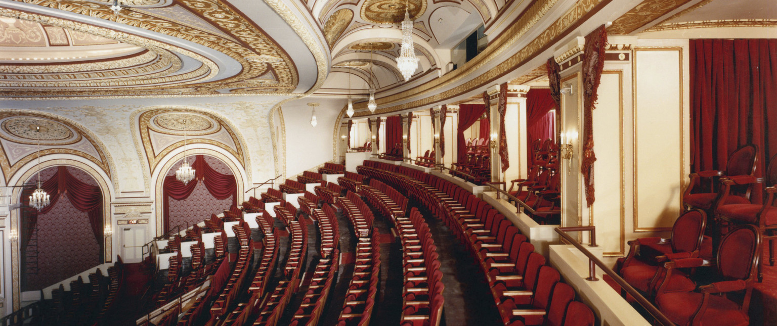 Box seats and back rows of audience in the Connor Palace Theater, with red seats and white walls with gold scroll details