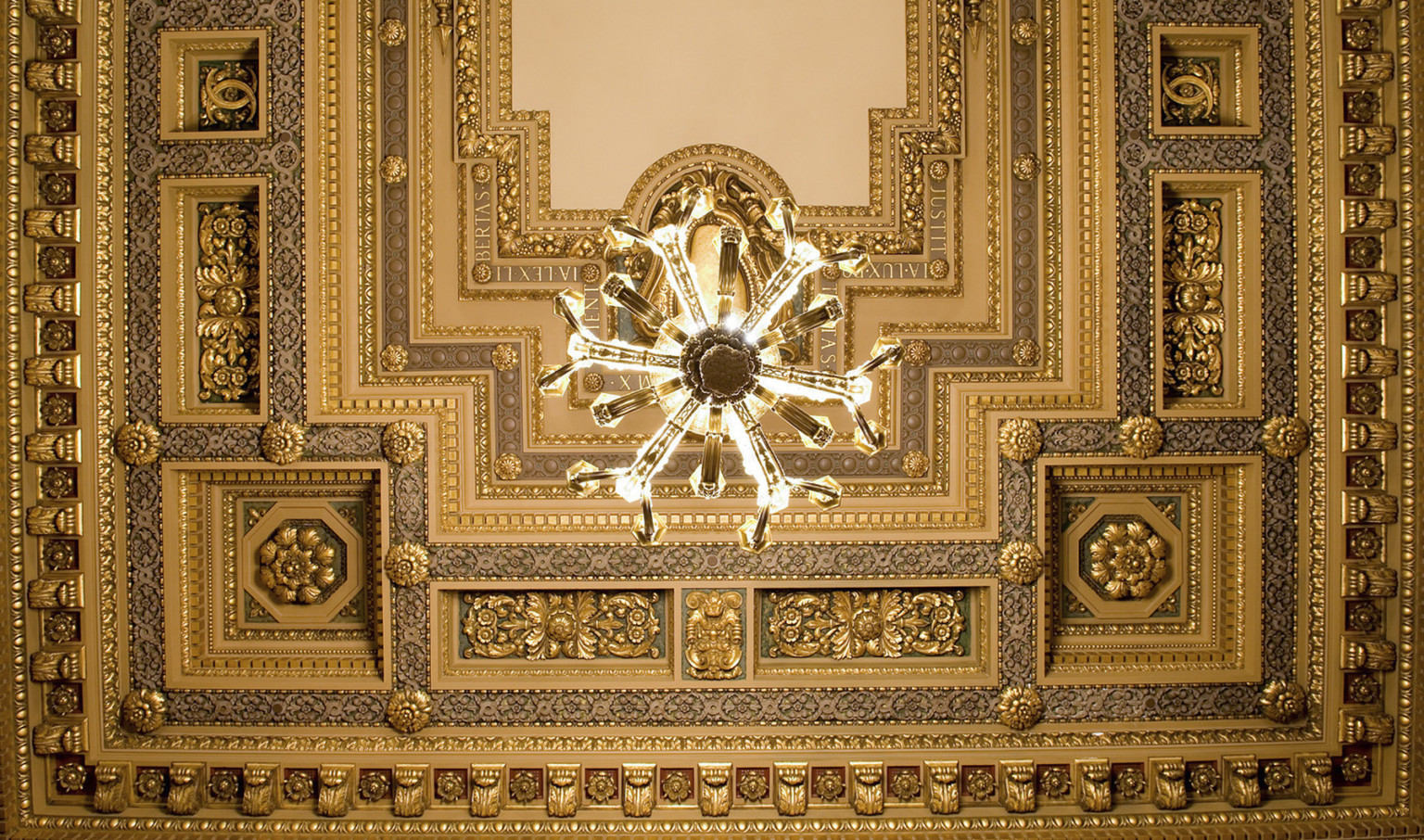Looking up at ornate orthogonal ceiling detailed with floral medallions, roman phrases, cornice edging, and offset chandelier