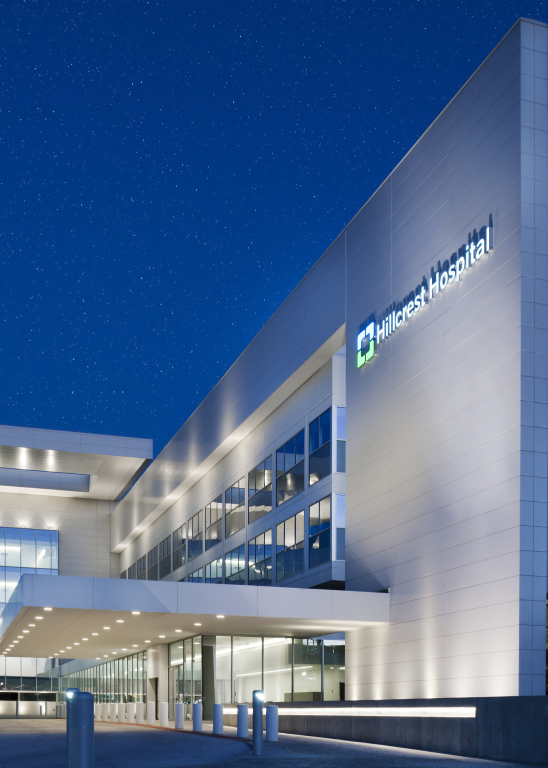 Silver building entry with floor to ceiling windows illuminated at night. Lit sign on upper right reads Hillcrest Hospital