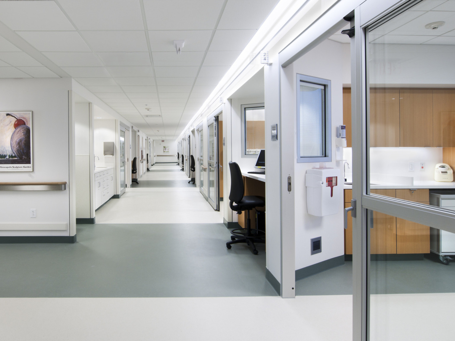 White hallway with patient rooms, with a desk between every other room. Grey stripes run across the width of the hallway