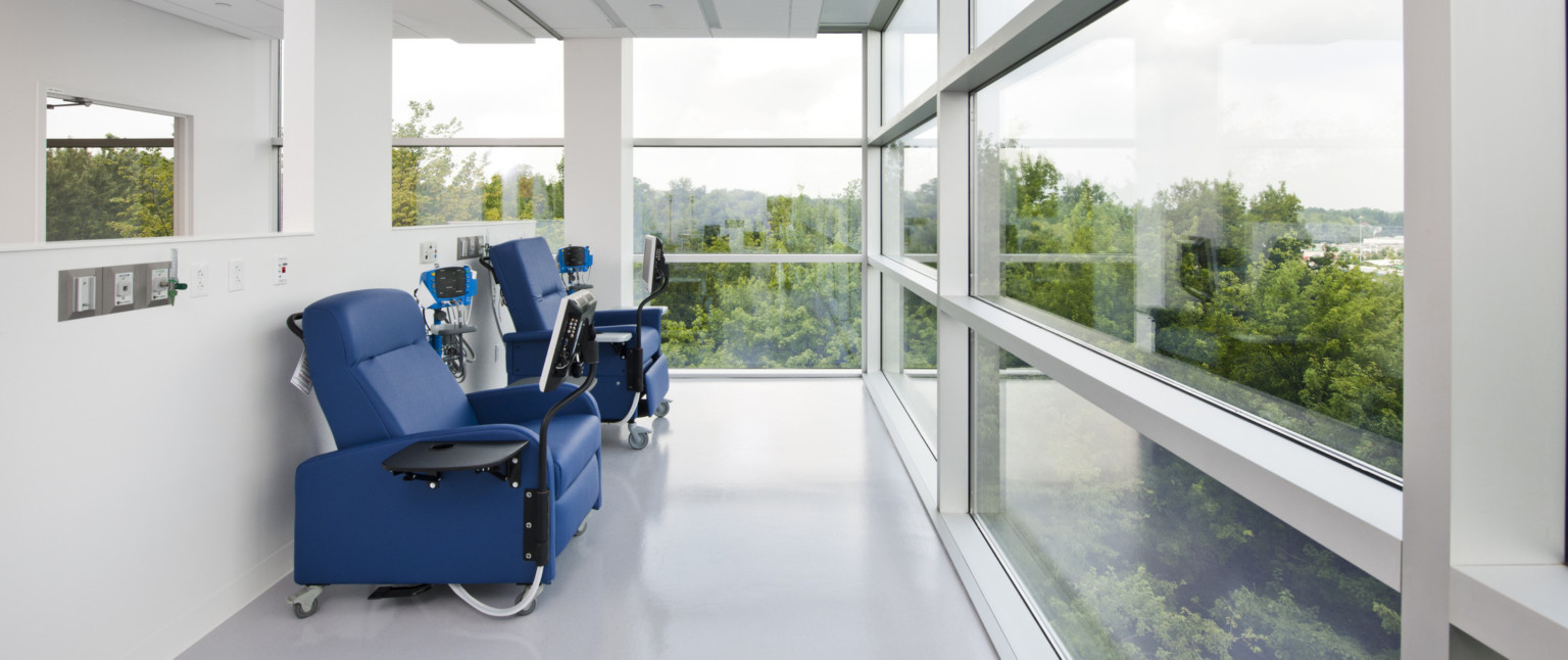 Blue chairs with attached table and screens next to half wall. Chairs face floor to ceiling window with view of trees