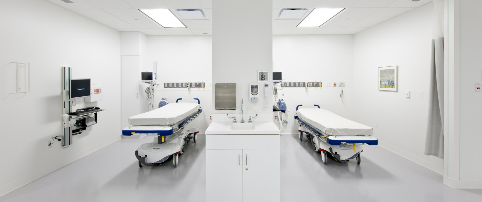 Two hospital beds in white room with equipment left. Desk and computer extend from left wall. White pillar center with faucet