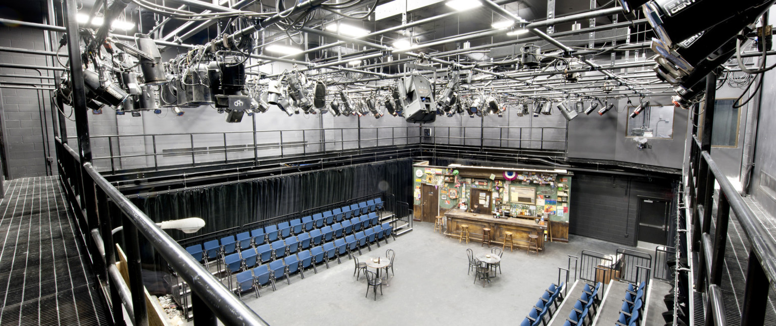 Black box theater with side seating, lights hang on a metal grid above. The stage is set with tables, chairs, and a bar